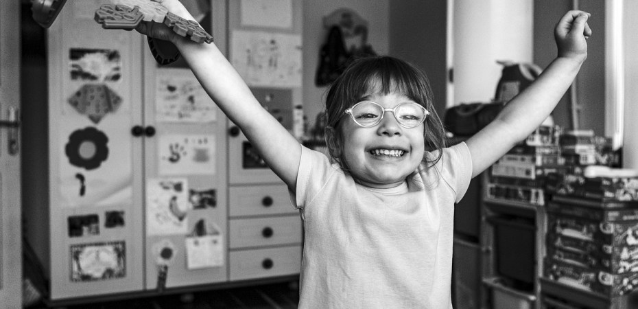 Schwarz- weiß Foto: Ein kleines Mädchen mit Brille reißt vor Freude die Arme in die Luft
