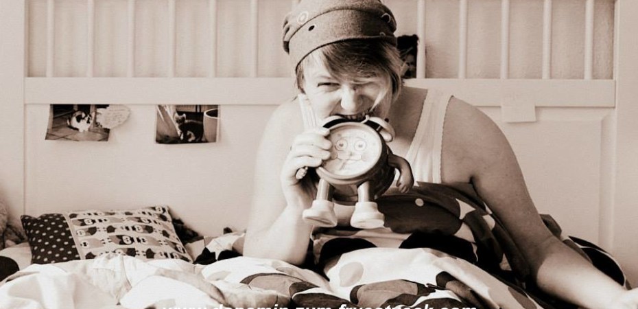 Frau mit Mütze im Bett. Sie sitzt im Bett und beißt in einen Wekcer