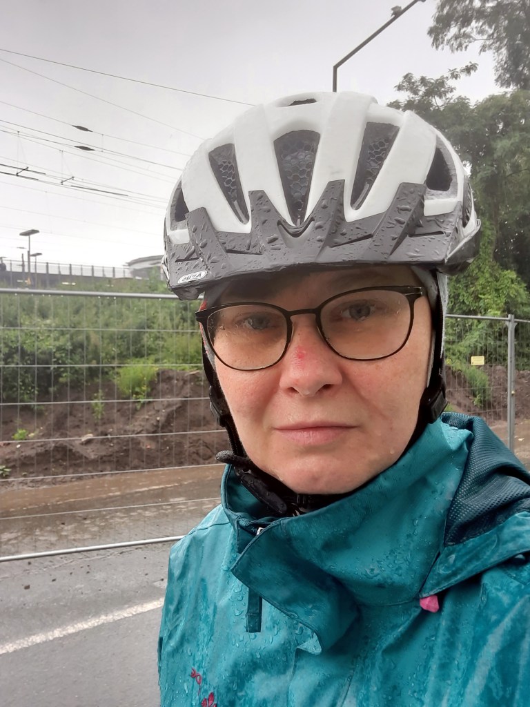 Eine Frau mit Brille, weißem Fahrradhelm und blauer Regenjacke, die vom Regen nass ist
