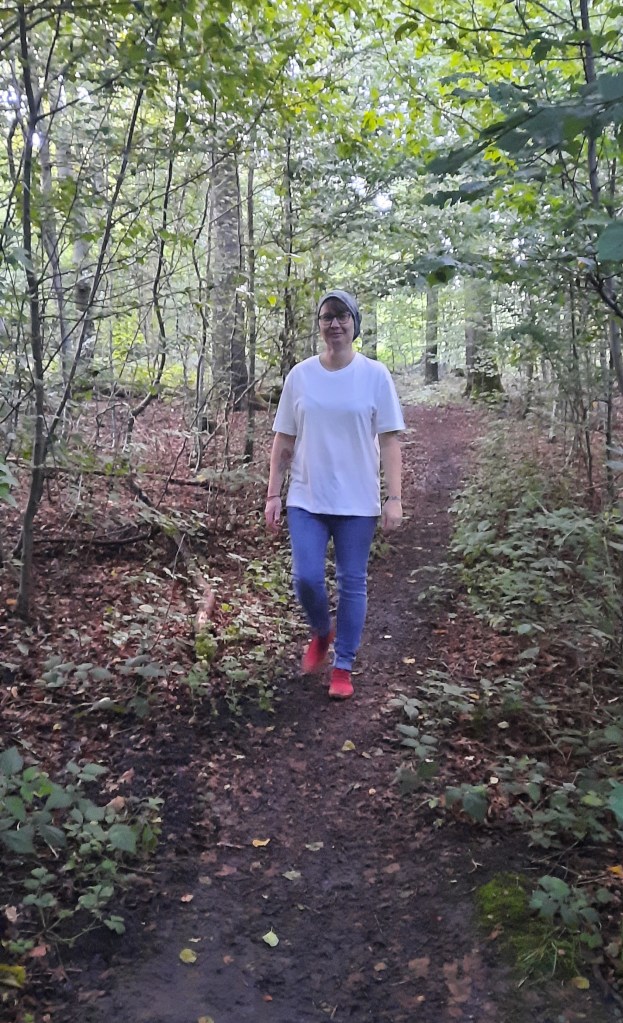 Eine Frau mit Mütze, Brille, weißen T-Shirt, blauer Jeans und roten Schuhen, die im Wald spazieren geht