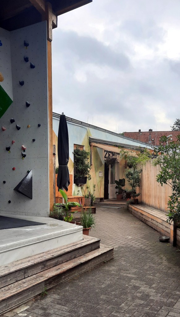 Links sieht man ein paar Boulder an einer Außenwand. Rechts führt ein Weg in den Eingangsbereich der Boulderhalle