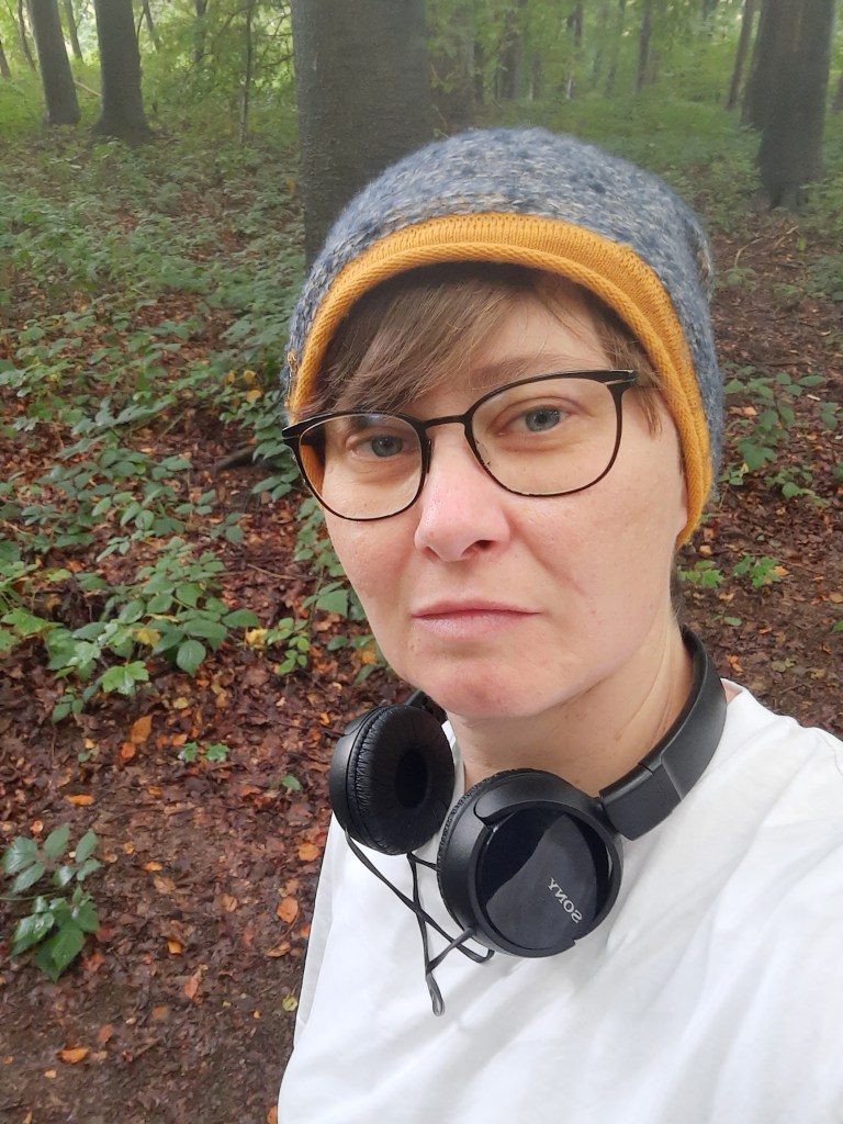 Eine Frau, die eine schwarze Brille und eine grau-orangene Mütze trägt. Um den Hals trägt sie Kopfhörer. Sie geht im Wald spazieren und macht ein Selfie.