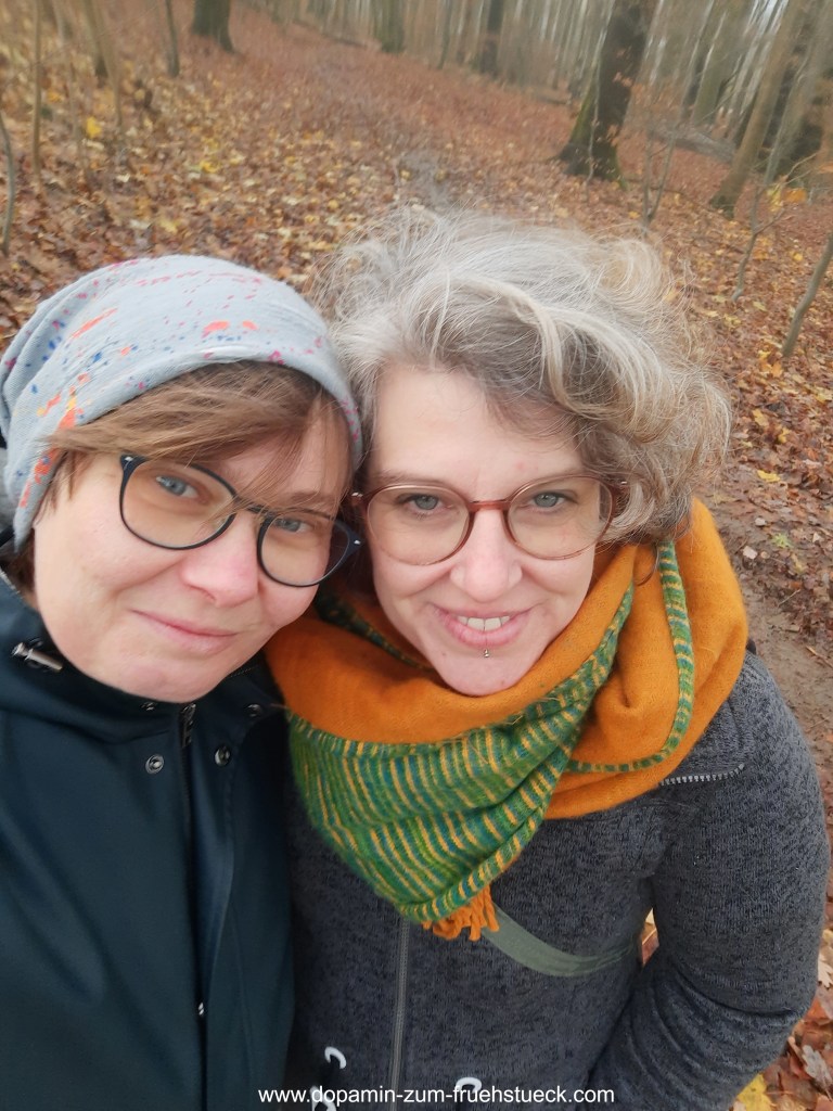 Zwei Frau mit Brille, die im Wald ein Selfie machen. Frau links trägt eine graue Mütze und eine grüne Jacke, Frau rechts hat graue Haare und trägt einen orange-grünen Schal und eine graue Jacke