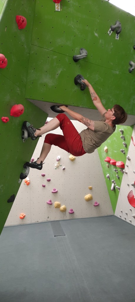 Frau mit kurzen Haaren, roter Hose und grauem T-Shirt an einer Boulderwand