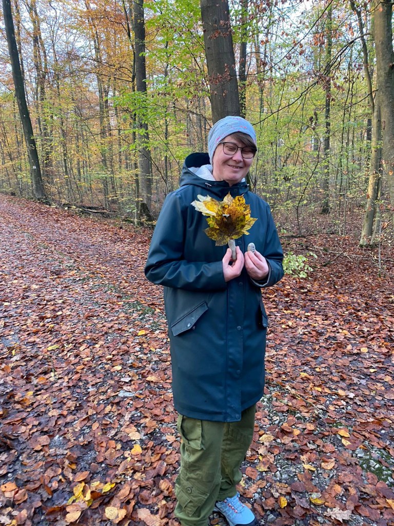 Eine Frau mit Brille, grauer Mütze, grüner Regenjacke und grüner Hose, die einen Strauß bunter Blätter in der Hand hält