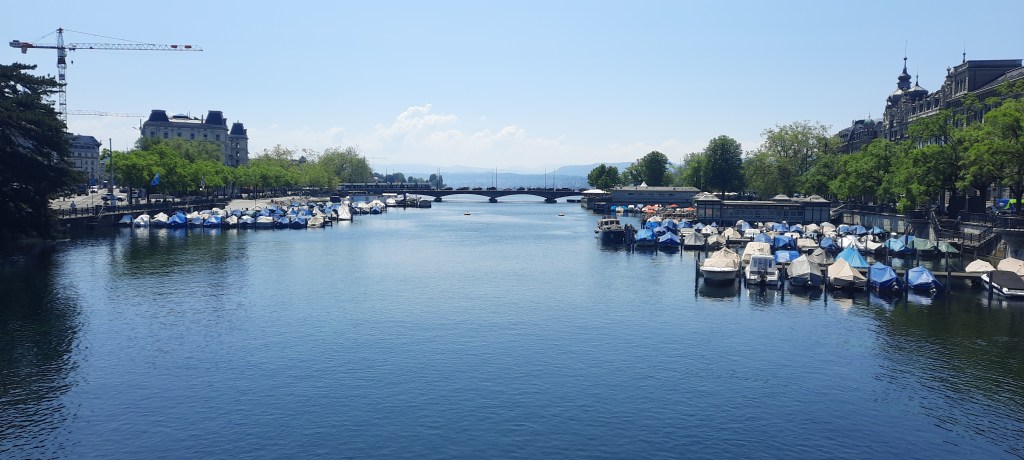 Zürichsee- blauer See mit einer Brücke im Hintergrund. Im Vordergrund sind viele kleine Boote links und rechts zu sehen