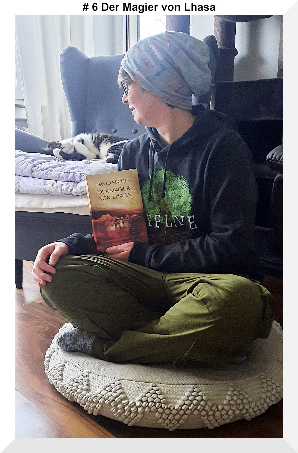 Eine Frau mit Mütze auf einem Sitzkissen, die ein Buch in der Hand hält. Sie blickt hinter sich auf den Sessel, denn dort liegt ein schwarz- weißer Kater