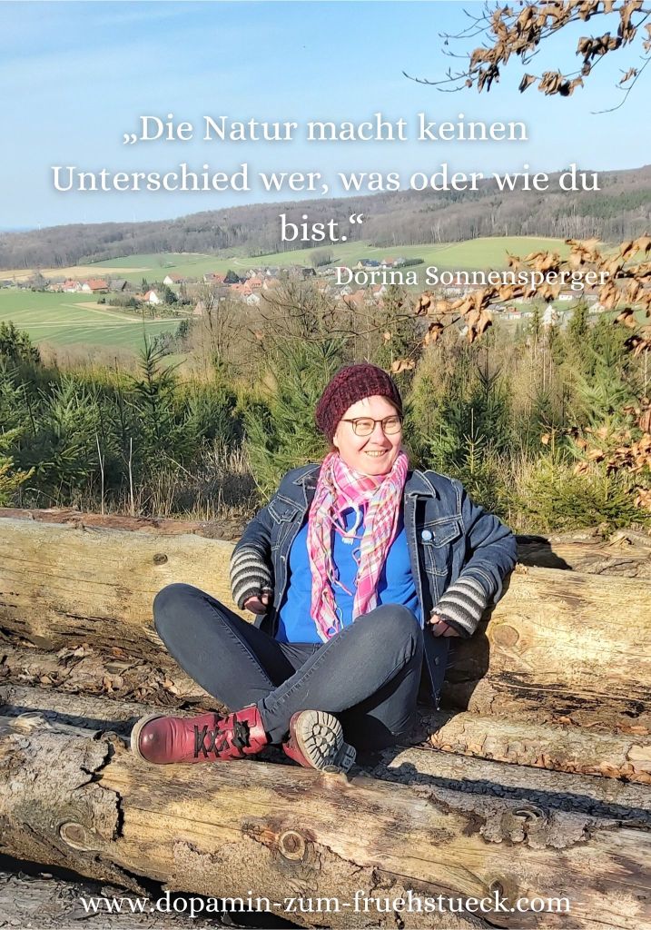 Eine Frau mit roter Mütze und Brille, die auf gefällten Baumstämmen sitzt. Der Himmel ist blau und im Hintergrund kann man einzelne Häuser sehen.