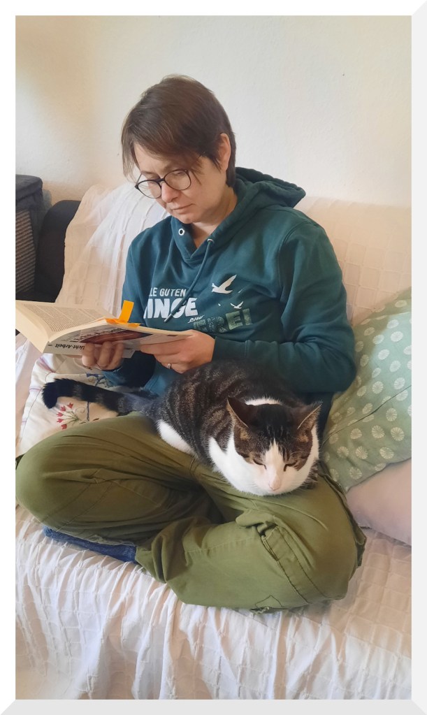 Eine Frau mit kurzen Haaren,  Brille, grünem Hoodie und grüner Hose sitzt auf einem Sofa und liest. Auf ihrem Schoß liegt eine schwarz- weiße Katze.