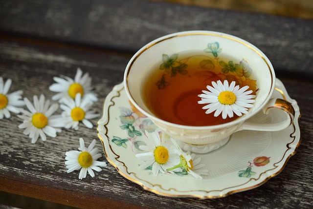 Eine Tasse Tee. In dem Tee schwimmt eine Kamillenblüte und neben der Tasse liegen links fünf weitere Kamillenblüten