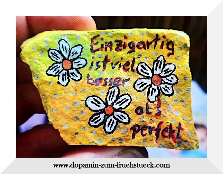 Ein bemalter Stein. Die Hintergrundfarbe ist gelb und auf dem Stein sind drei Gänseblümchen gemalt und in rot- blauer Schrift steht geschrieben: Einzigartig ist besser als perfekt.
