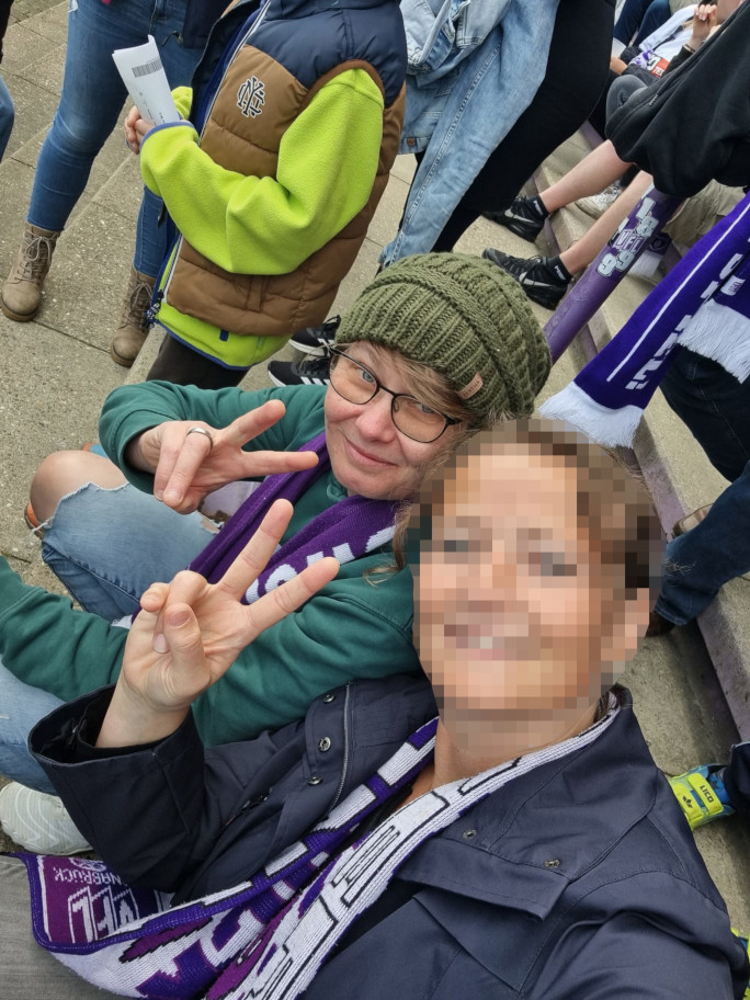 Zwei Frau im Fussballstadion. Beide schauen in die Kamera und zeigen ein Peace- Zeichen. Eine Frau trägt eine Grüne Mütze und Brille, die zweite Frau ist verpixelt.