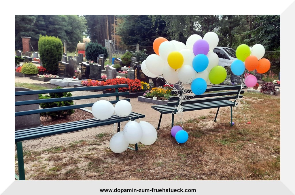 Auf einem Friedhof stehen zwei grüne Bänke, an denen viele bunte Luftballons befestigt sind