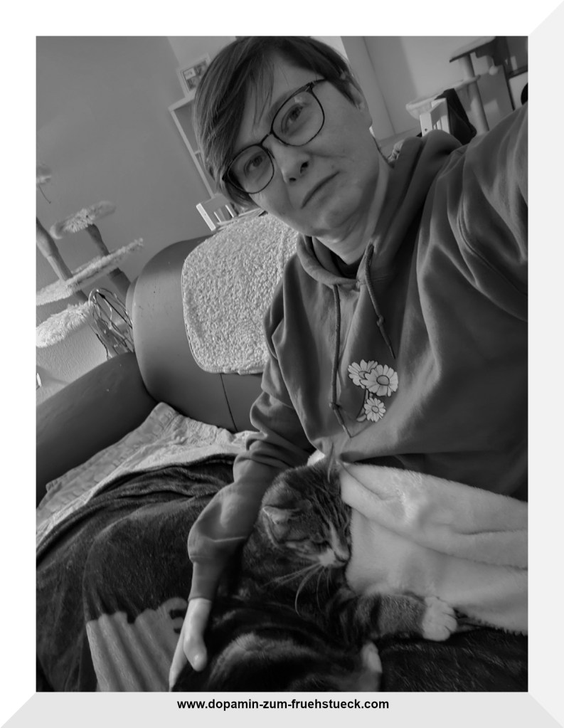 Schwarz-Weiß Foto einer Frau mit kurzen Haaren und Brille, die auf einem Sofa sitzt. In ihrem Schoß liegt eingerollt eine Katze.