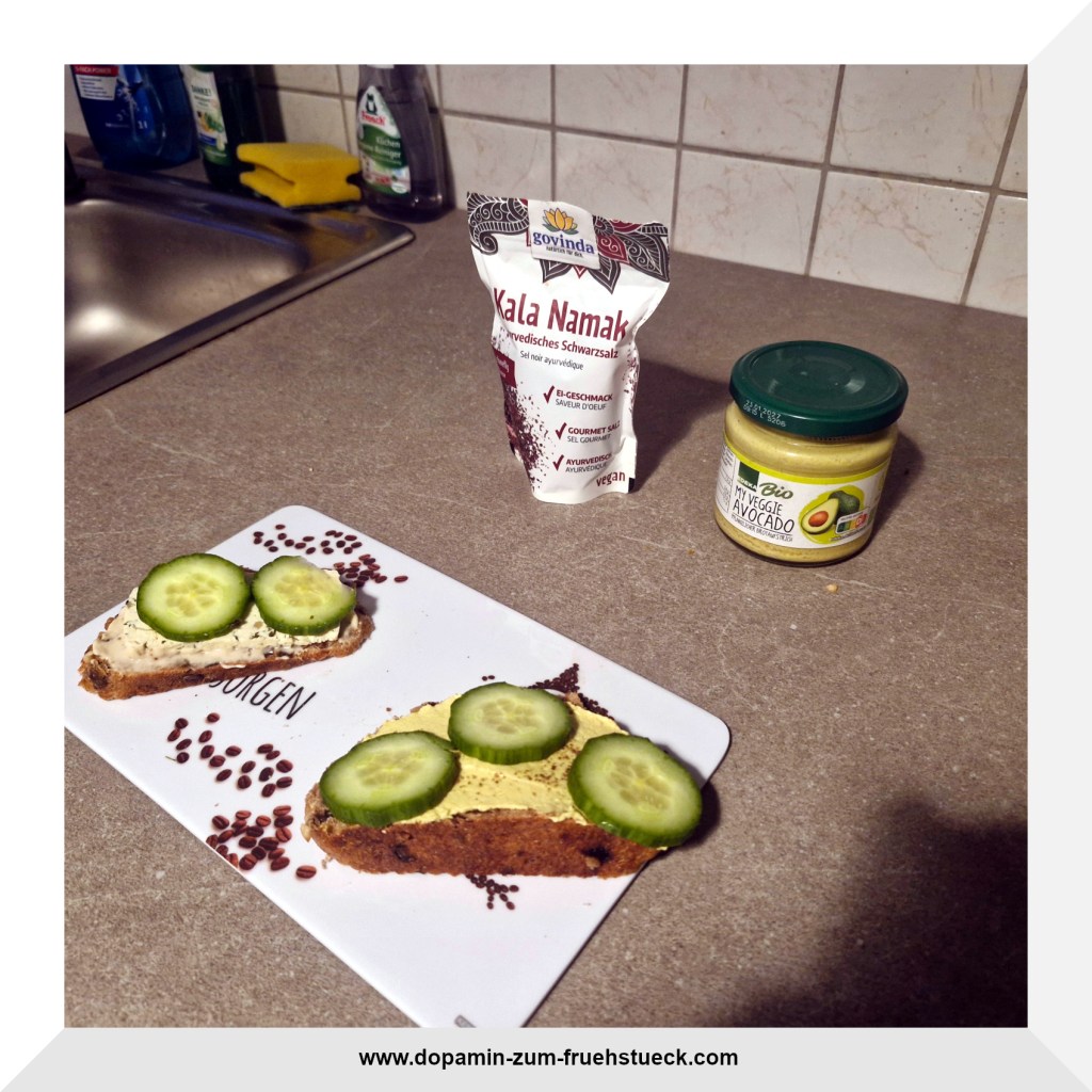 Zwei Brote beschmiert mit Aufstrich, Tofu und Gurke auf einem Brett. Daneben Kala Namak und Aufstrich