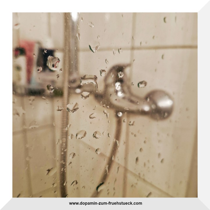 Wassertropfen auf einer Duschwand; im Hintergrund verschwommen zu sehen der Wasserhahn