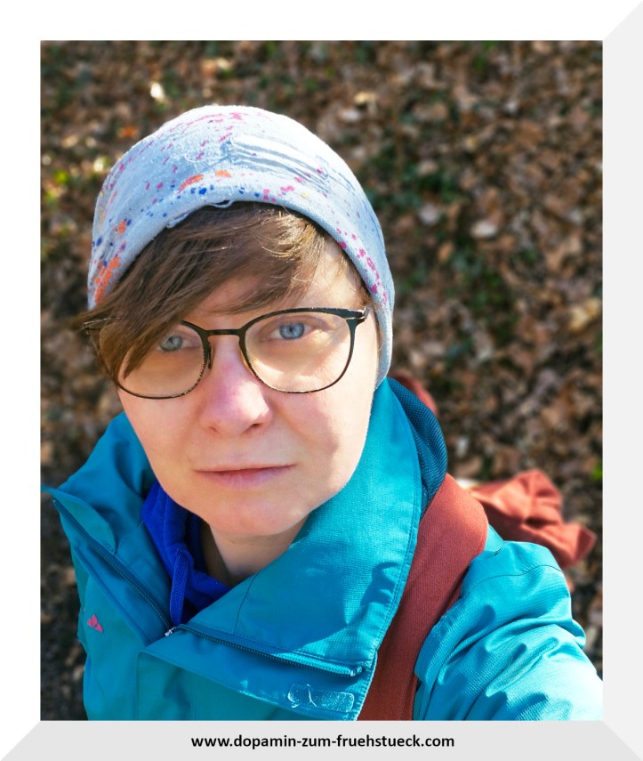 Eine Frau mit Brille, Mütze und blauer Jacke, die im Wald steht und ein Selfie von oben (aus der Vogelperspektive) macht