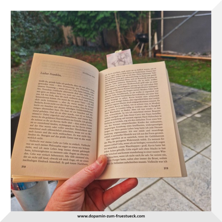 Ein aufgeschlagenes Buch in einer Hand. Das Foto wurde draußen gemacht, da man im Hintergrund die Terrasse erahnen kann