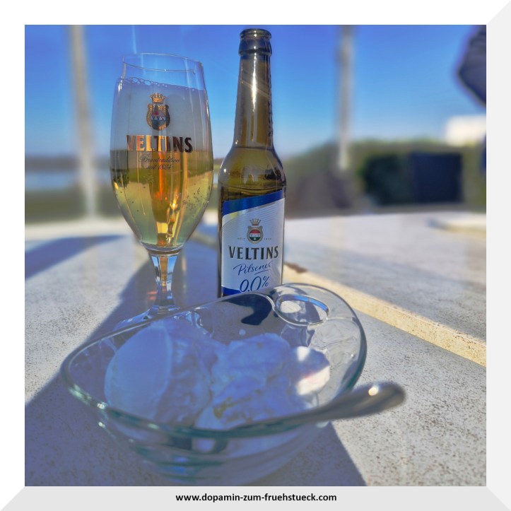 ein Glas und eine Flasche alkoholfreies Bier und eine Schüssel mit zwei Kugel Eis auf einem Tisch. Im Hintergrund kann man Wasser erkennen