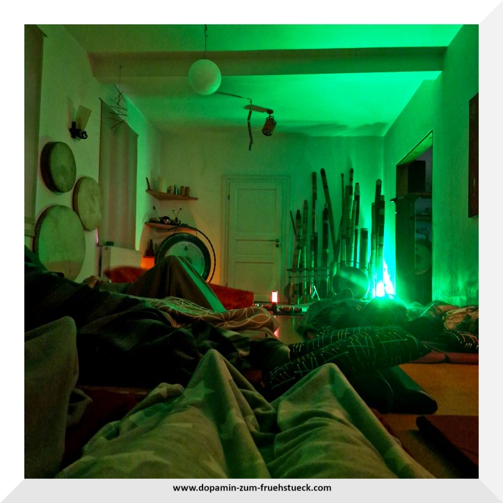 Ein abgedunkelter Raum, in dem nur ein kleines grünes Licht brennt. Auf dem Boden liegen in Decken eingekuschelt 9 Menschen. Im Hintergrund kann man Didgeridoos, Klangschalen und Trommeln stehen sehen