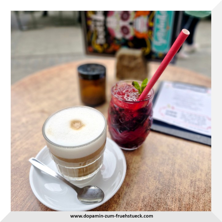 Ein Glas mit Kaffee und Milchschaum und daneben eine rote Limonade. Beides steht auf einem Tisch- im Hintergrund kann man verschwommen eine Speise-/Getränkekarte erkennen