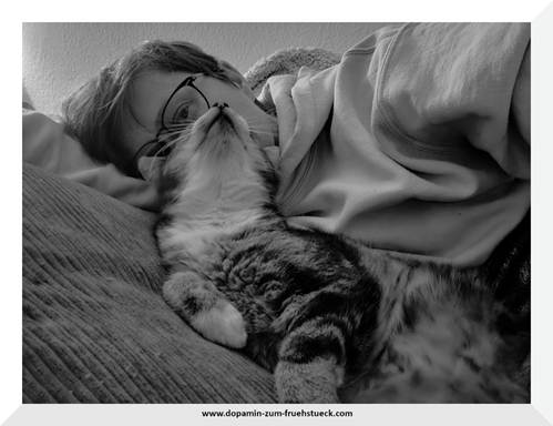 Eine Frau und eine Katze, die auf einem Bett oder Sofa liegen und kuscheln. Man erkennt nur einen Teil des Kopfes der Frau, weil die Katze sich direkt an den Kopf der Frau lehnt