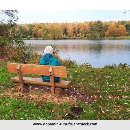 Ein Mensch mit Mütze auf einer Bank am See. Man sieht den Menschen nur von hinten